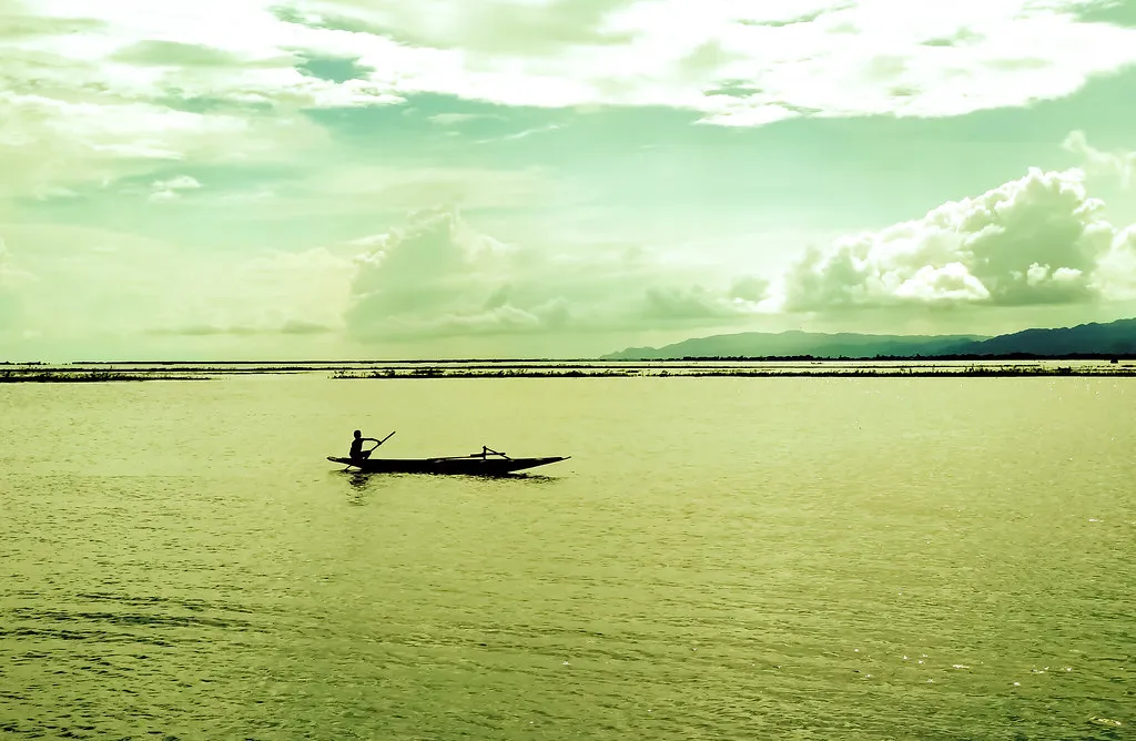 Lone boatman paddling across a wide haor wetland under a pale sky